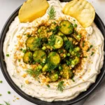 A bowl of creamy dill pickle dip topped with sliced pickles and fresh dill, served with potato chips.