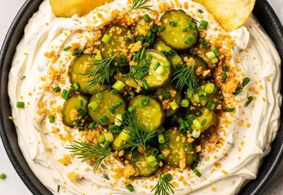 A bowl of creamy dill pickle dip topped with sliced pickles and fresh dill, served with potato chips.