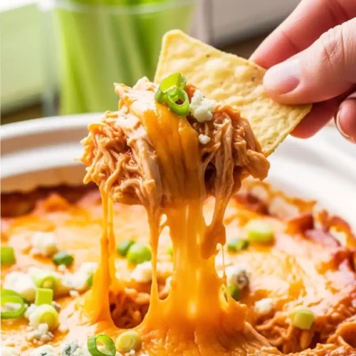 A hand scooping a generous portion of creamy buffalo chicken dip with a tortilla chip, showing a long cheese pull and toppings of green onions and blue cheese.