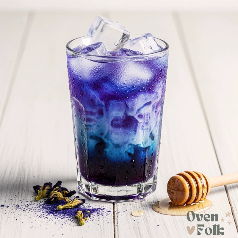 A tall glass of iced butterfly pea flower latte with a blue and white gradient, condensation on the glass, and a honey dipper on a white wooden table.
