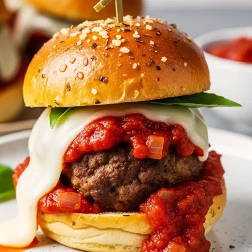 Close-up of a meatball slider with melted mozzarella, marinara sauce, and fresh basil on a garlic butter bun, secured with a bamboo skewer.