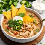 A close-up of creamy white chicken chili in a speckled white bowl, topped with jalapeños, sour cream, and cilantro, served with tortilla chips on a wooden board.