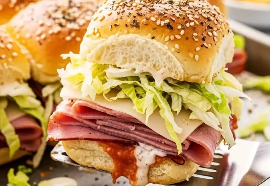 A spatula lifting a baked Italian slider with salami, cheese, and lettuce from a sheet pan.