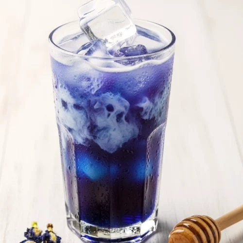 A tall glass of iced butterfly pea flower latte with a blue and white gradient, topped with dried purple flowers and lavender honey on a white wooden table.