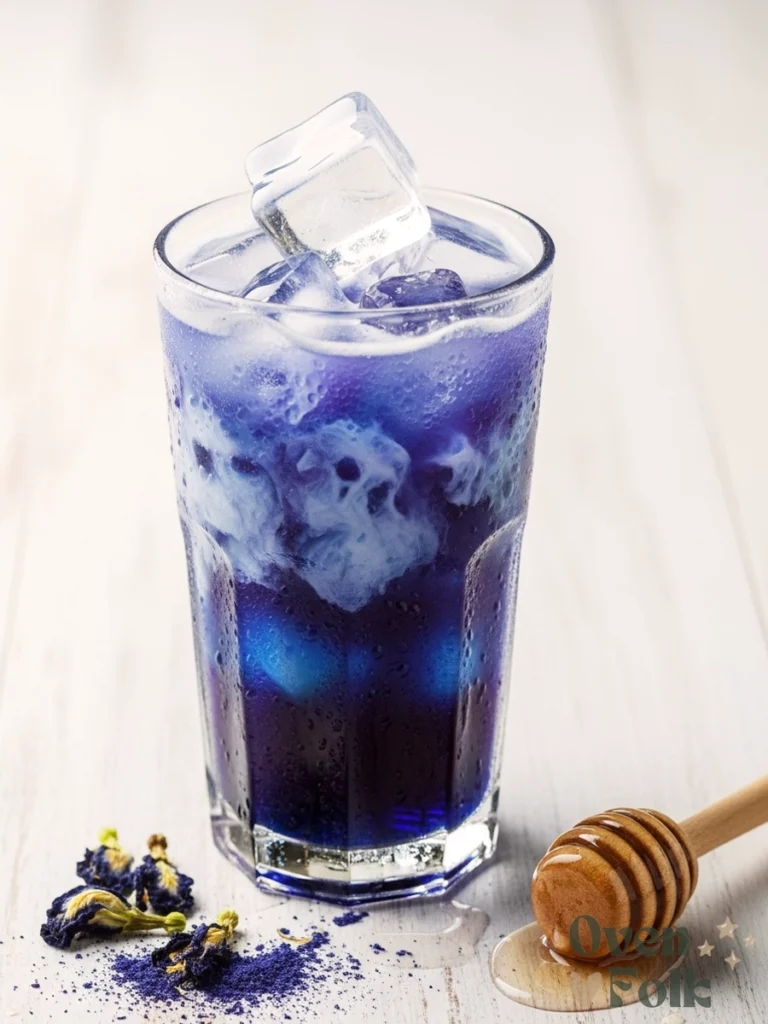 A tall glass of iced butterfly pea flower latte with a blue and white gradient, topped with dried purple flowers and lavender honey on a white wooden table.