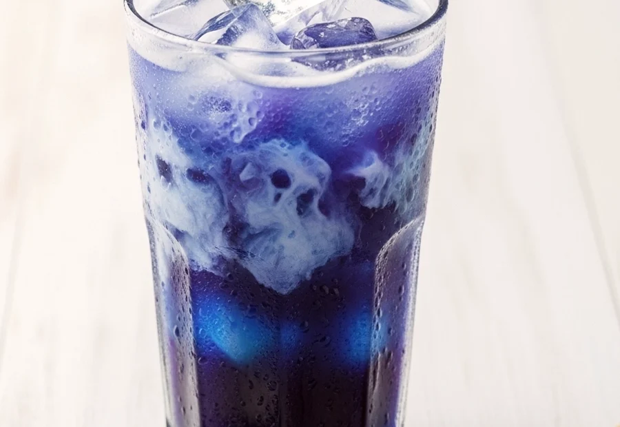 A tall glass of iced butterfly pea flower latte with a blue and white gradient, topped with dried purple flowers and lavender honey on a white wooden table.