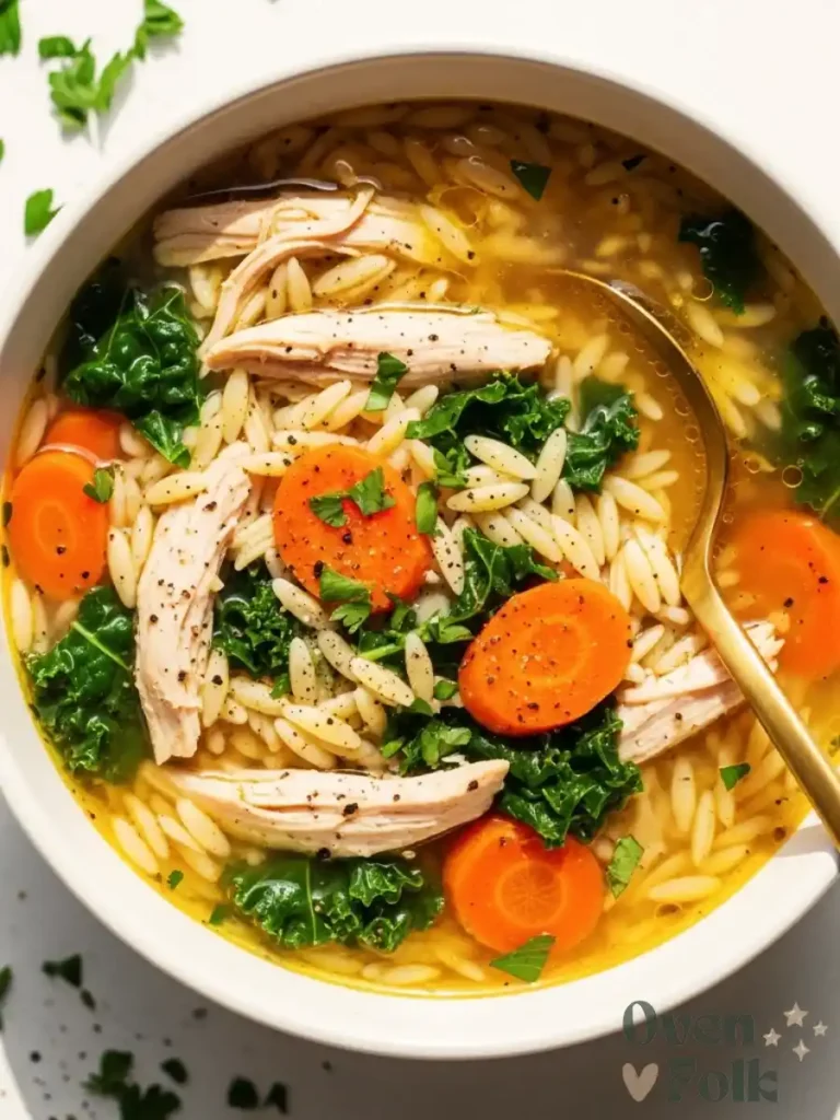 Top-down view of a white bowl filled with lemon chicken orzo soup, carrots, kale, and a gold spoon.
