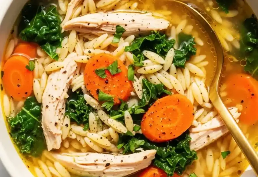 Top-down view of a white bowl filled with lemon chicken orzo soup, carrots, kale, and a gold spoon.