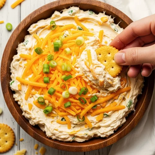 Top down view of a wooden bowl filled with creamy Million Dollar Dip loaded with shredded cheddar cheese, and green onions, being scooped with a round cracker.