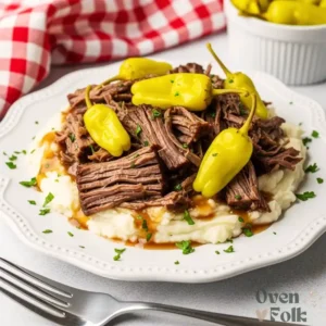 A close up view of shredded beef pot roast with brown gravy and yellow pepperoncini peppers served on top of creamy mashed potatoes on a white plate.