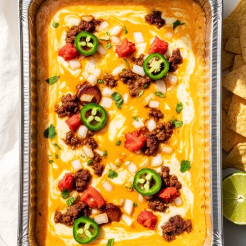 Overhead view of bubbling smoked queso dip in an aluminum tray with melted cheese, chorizo, and jalapeños on a white marble surface.