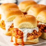 Shredded buffalo chicken sliders with creamy ranch sauce dripping over the side onto parchment paper.