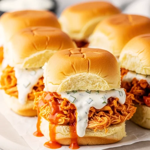 Shredded buffalo chicken sliders with creamy ranch sauce dripping over the side onto parchment paper.